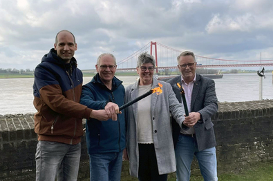 Tim Terhorst (Stadt Emmerich am Rhein) und die niederländischen Organisatoren Ton Masselink, Christa van Dee und Henk Groote mit zwei Fackeln vor der Emmericher Rheinbrücke.