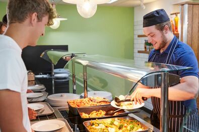 Mitarbeiter eines Caterer bedient einen älteren Schüler in der Schulmensa.